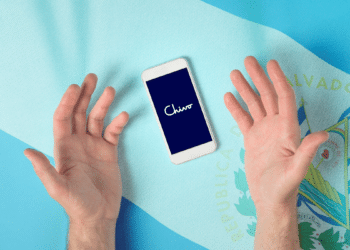 Manos reclamando a un teléfono con logo de Chivo Wallet y bandera de El Salvador de fondo.