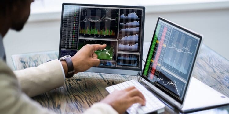 Un hombre frente a dos pantallas de computador con gráficos de precios