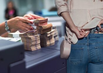 mujer sostiene arma escondida dentro de pantalon mientras cejro de banco apila dinero sobre el mostrador