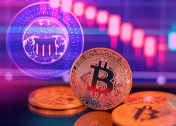 moneda de bitcoin en frente de pantalla con gráfico bajista y logo de la Reserva Federal
