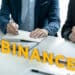 Logo de Binance en mesa con personas, documentos y maso de juez llegando a un acuerdo