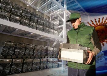 hombre sosteniendo equipo asic para la mienería de bitcoin y criptomonedas junto a estantes llenos de quipos asic con bandera de argentina en el fondo