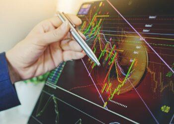 mao de trader sosteniendo boligrafo sobre pantalla, señalando bajada en grafico de precio con moneda de bitcoin en el fondo