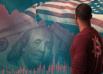 hombre con logo de bitcoin en camisa obsercando grafico descendente con billete de dolar y bandera de los Estados Unidos en el fondo
