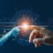Una mano mecanica hace contacto con una mano humana generando un chispa luminosa.