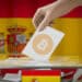 persona depositando papel de voto con logo de Bitcoin en cajeta de votaciones con bandera de España en el fondo