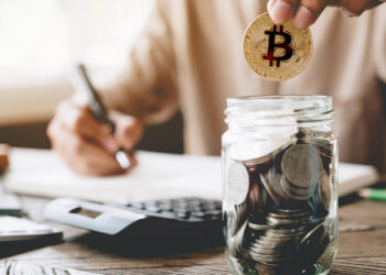 persona colocando moneda de bitcoin en frasco con monedas y haciendo cuentas en el fondo