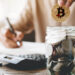persona colocando moneda de bitcoin en frasco con monedas y haciendo cuentas en el fondo