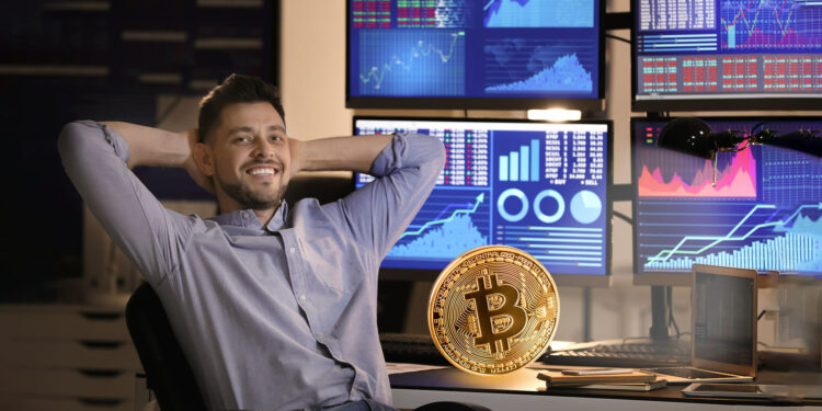 Un trader joven sentado frente a varias pantallas con graficos de precios de activos y una moneda de bitcoin a su lado.