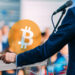 logo de Bitcoin sobre mesa con hombre dando discurso ante un publico