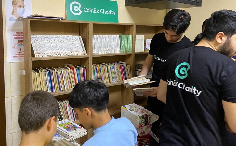 Varios estudiantes con franelas negras de CoinEx Charity revisando libros al lado de estante lleno de publicaciones.