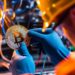 un trabajador con ropa de minero sostiene una moneda de bitcoin, frente a máquinas y cablados
