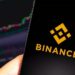 Teléfono móvil con logo de Binance en la pantalla y un gráfico de velas al fondo en un ordenador.