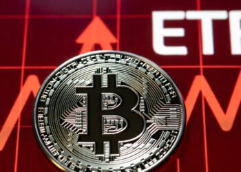Moneda física de bitcoin en primer plano, detrás un gráfico financiero en rojo intenso y las letras ETF.