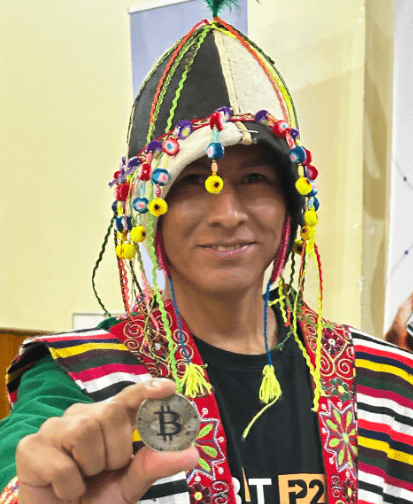 Una persona con traje típico de Bolivia luce una moneda física de bitcoin.