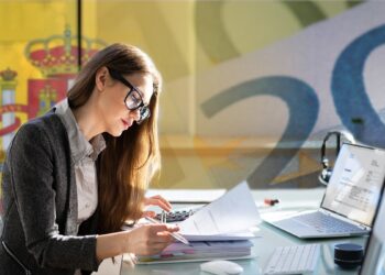 Mujer joven de lentes revisa documentos en el escritorio.