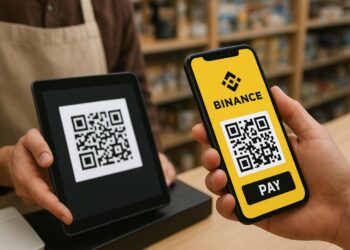 Una persona se encuentra en una tienda realizando un pago con su teléfono móvil. En la pantalla del celular se ve claramente el logotipo amarillo de Binance y un código QR con el botón “PAY” debajo. Frente a ella, un empleado sostiene una tableta negra que muestra otro código QR para recibir el pago. Ambos están en un mostrador de madera, con estantes llenos de productos desenfocados al fondo. La iluminación es cálida y natural, dando un ambiente acogedor y cotidiano, típico de una tienda moderna.