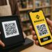 Una persona se encuentra en una tienda realizando un pago con su teléfono móvil. En la pantalla del celular se ve claramente el logotipo amarillo de Binance y un código QR con el botón “PAY” debajo. Frente a ella, un empleado sostiene una tableta negra que muestra otro código QR para recibir el pago. Ambos están en un mostrador de madera, con estantes llenos de productos desenfocados al fondo. La iluminación es cálida y natural, dando un ambiente acogedor y cotidiano, típico de una tienda moderna.