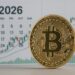 Moneda de bitcoin y en el fondo un calendario del año 2026 con un gráfico de velas verdes que simboliza la expectativa alcista.