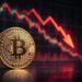 Moneda física de bitcoin dorada con gráficas en rojo de fondo.