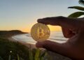 Una mano humana sosteniendo una moneda de bitcoin entre su dedo pulgar e índice y de fondo mar, sol y montañas de El Salvador.