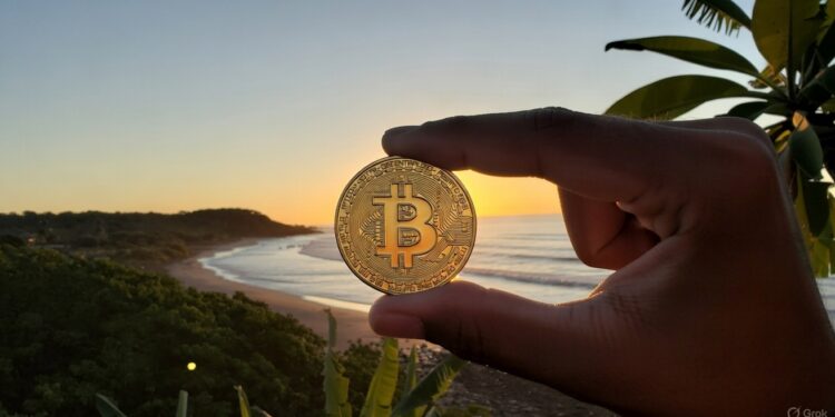 Una mano humana sosteniendo una moneda de bitcoin entre su dedo pulgar e índice y de fondo mar, sol y montañas de El Salvador.