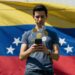 Una persona delante de una bandera de Venezuela sostiene un teléfono en sus manos y de él saltan monedas de USDT.