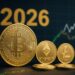 Monedas físicas doradas de bitcoin, ether y litecoin con un 2026 en el fondo y gráficas de mercado.